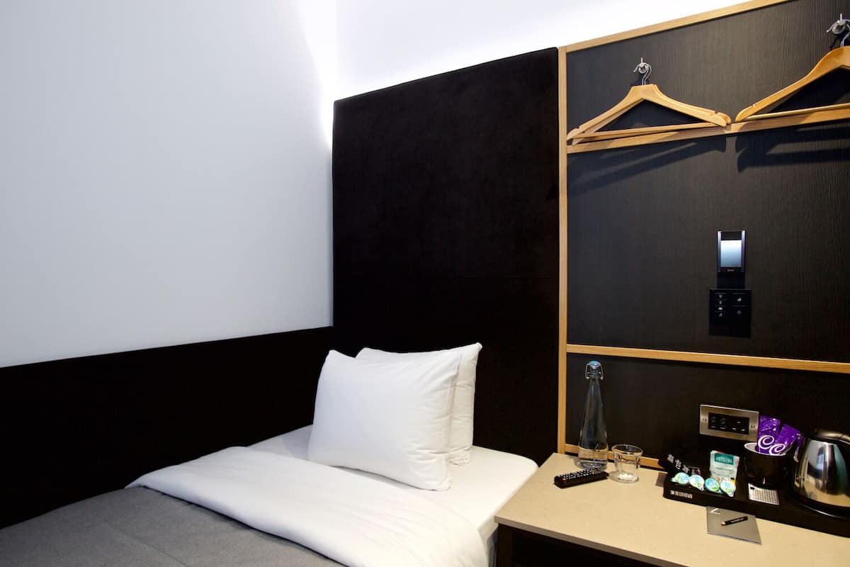 A hotel room with a bed, a bedside table with a remote control, a glass of water, and a tray with toiletries.
