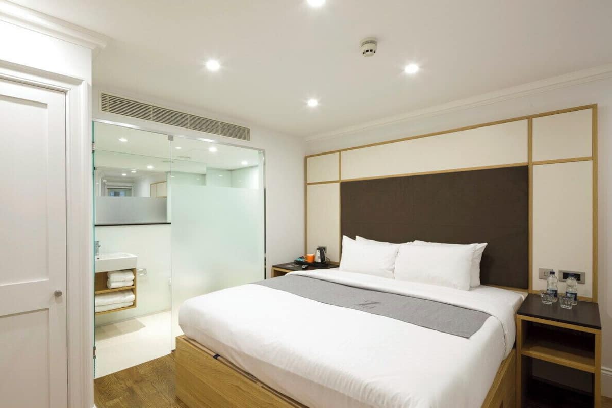 A modern hotel room with a large bed, a bedside table, a nightstand, a glass shower enclosure, and a white door.