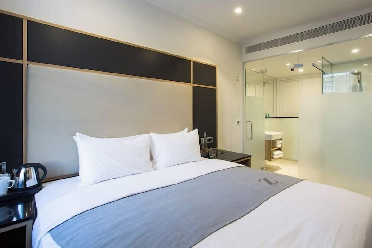A modern hotel room with a large bed, a bedside table with a kettle, and a glass shower enclosure.
