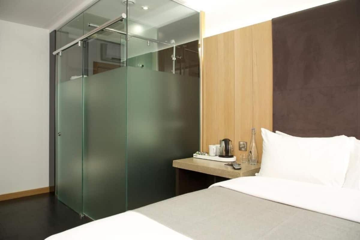 A modern hotel room with a bed, a small bedside table with a coffee maker, and a glass-enclosed shower area.