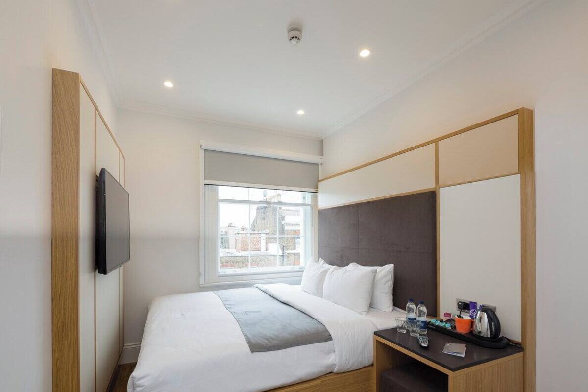 A modern hotel room with a wooden bed, a small desk with a chair, a television mounted on the wall, and a window with a view of buildings.