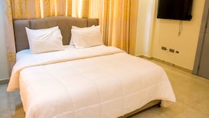 Een net opgemaakt bed met een witte deken en kussens, een flatscreen televisie aan de muur en een kamerbreed gordijn op de achtergrond.