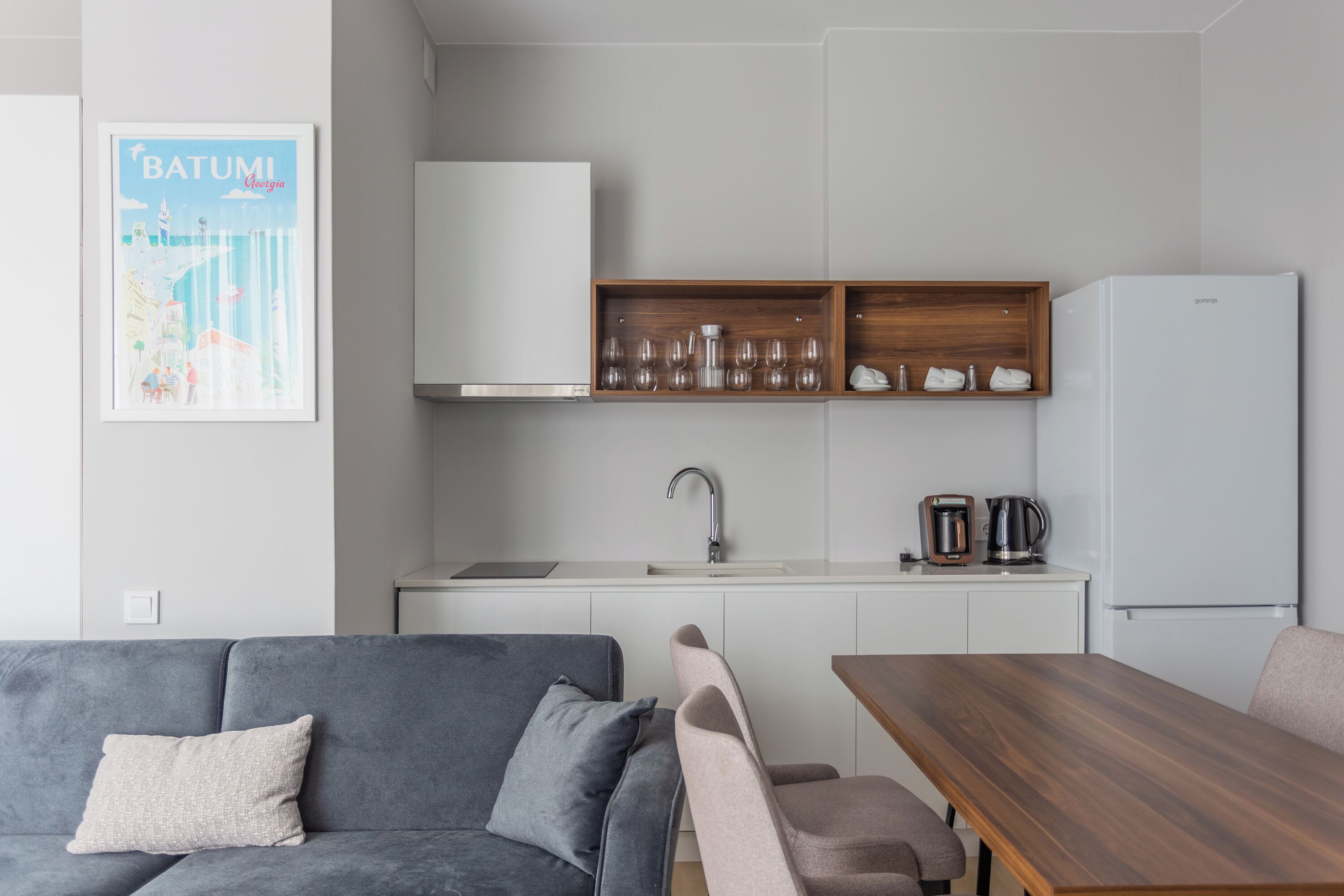 Eine moderne Küchenzeile mit einem weißen Kühlschrank, einer Holzauslage für Gläser und einem Wasserkocher sowie ein graues Sofa mit Kissen.