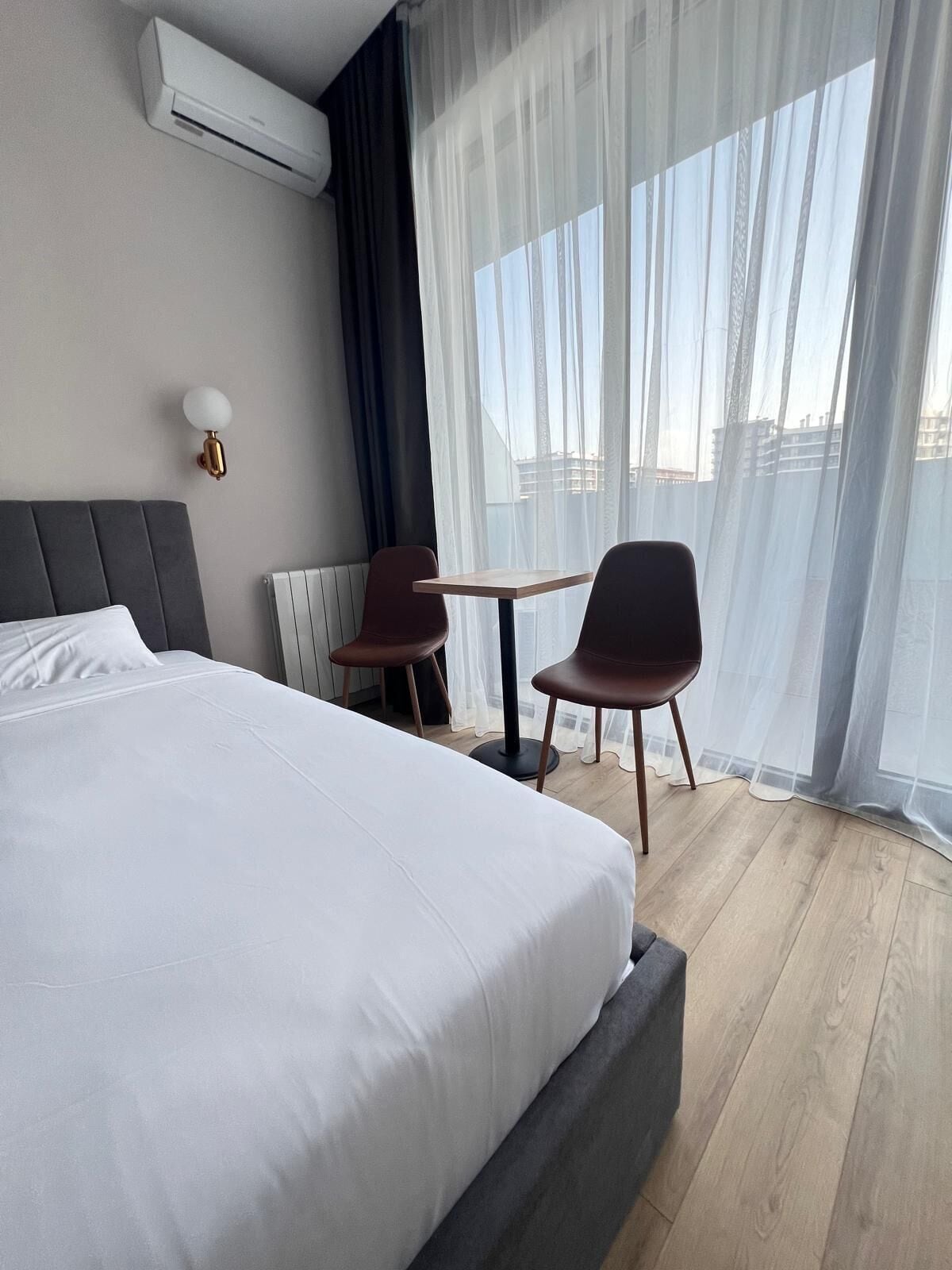Una camera d'albergo con un letto grande, un tavolino, due sedie, un condizionatore d'aria a muro e una finestra con tende leggere.