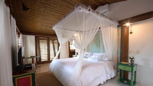 Une chambre à coucher avec un lit à baldaquin, un téléviseur posé sur un meuble, une petite table verte ornée d'une plante et un fauteuil en osier.