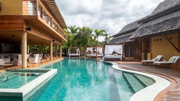Un espace piscine doté de chaises longues et d’un coin salon abrité, le tout entouré de bâtiments avec des balcons en bois.