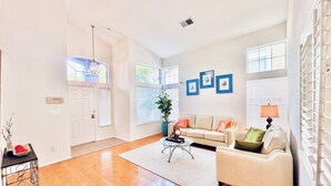 A spacious living room with hardwood floors, a beige sofa, a coffee table, and a potted plant. There are three framed pictures on the wall and a window with blinds.