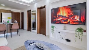 Ein modernes Wohnzimmer mit großem Fernseher, der ein Feuer zeigt, einer Essecke mit Stühlen und einer Küche im Hintergrund.