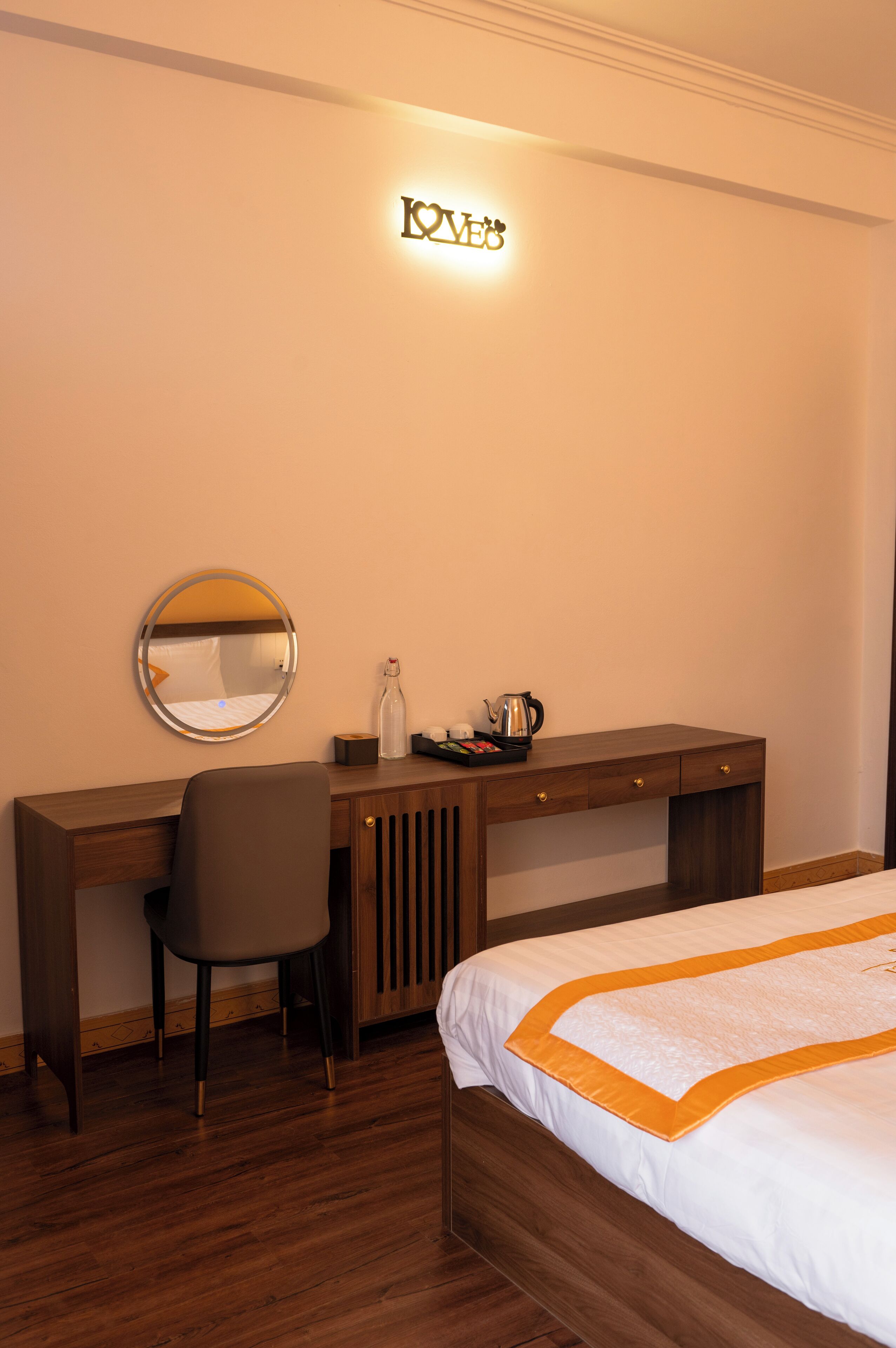 A hotel room with a wooden desk, a chair, a bed with an orange accent, a round mirror, and a wall-mounted light.