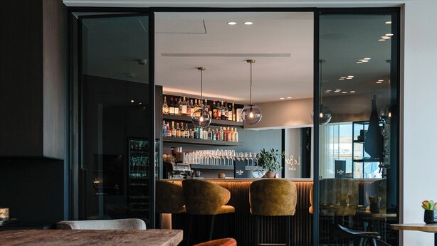 A modern bar area with a wooden bar counter, red upholstered chairs, and a glass partition separating the bar from the dining area. - Hotel du Bassin (Ostend)