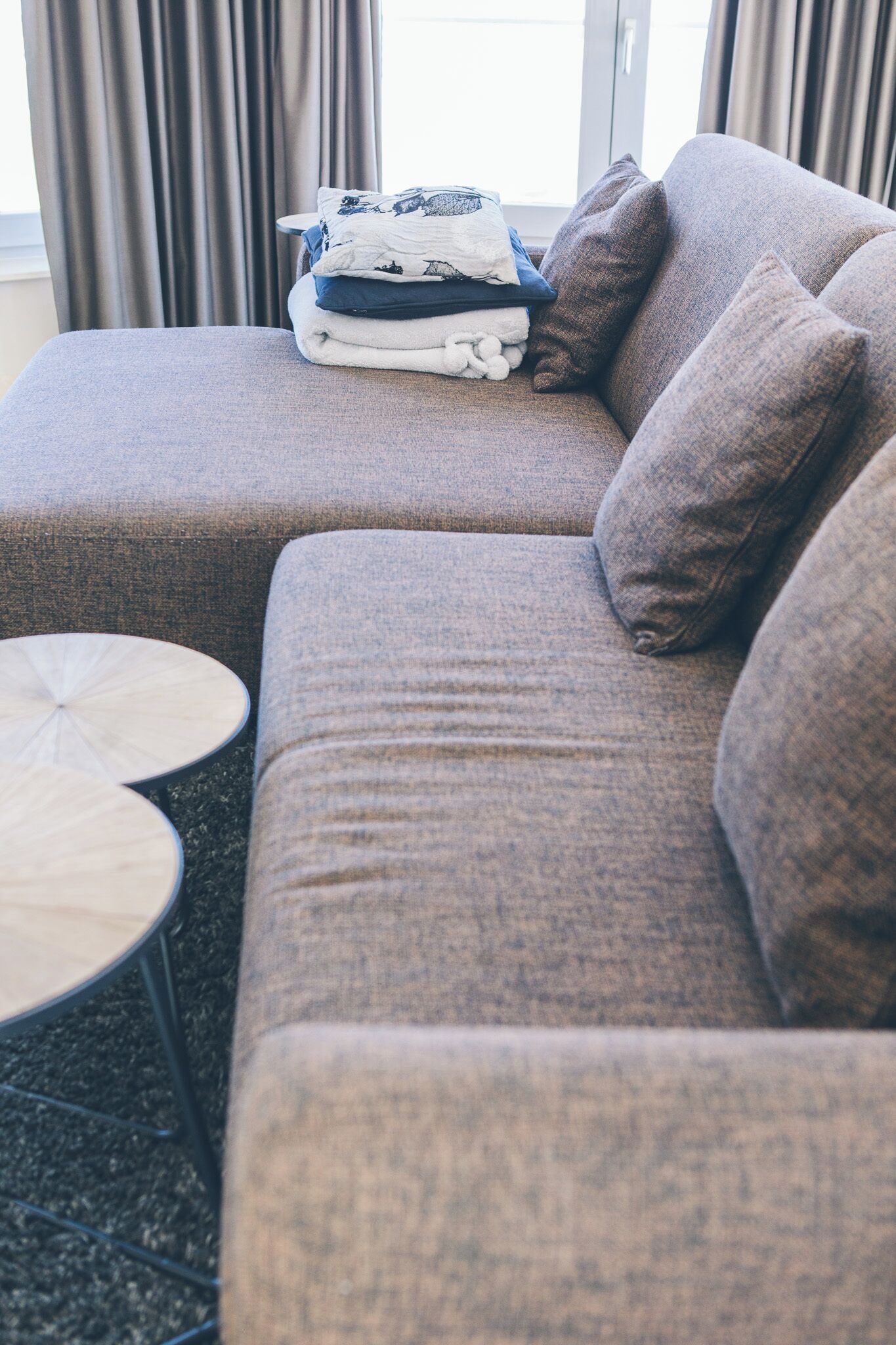 A modern living room with a grey sectional sofa, a small round coffee table, and a stack of folded blankets on the sofa.