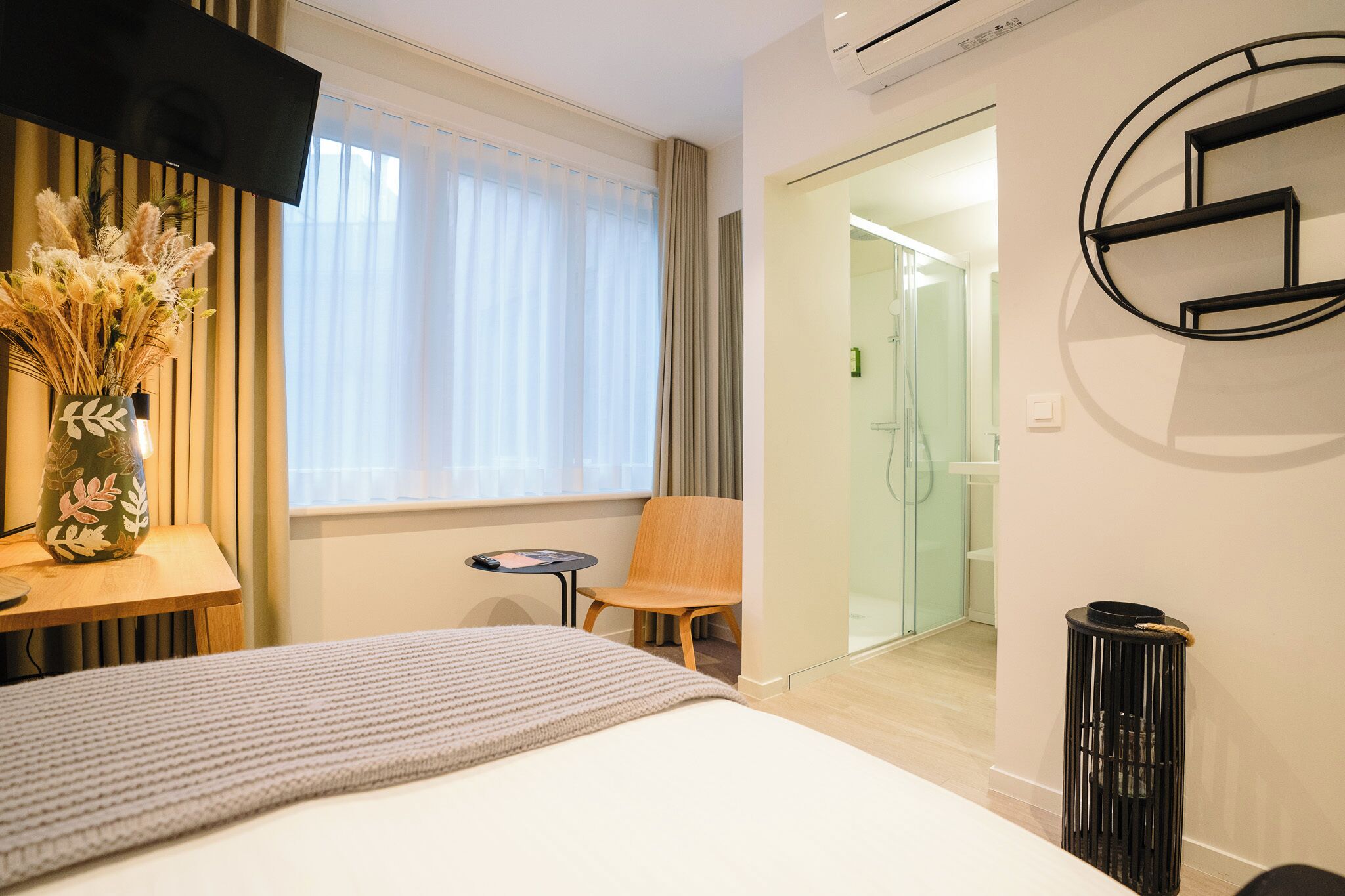 Una habitación de hotel moderna con una cama, una mesita con un florero de flores secas, una silla y una zona de ducha con mampara de cristal.