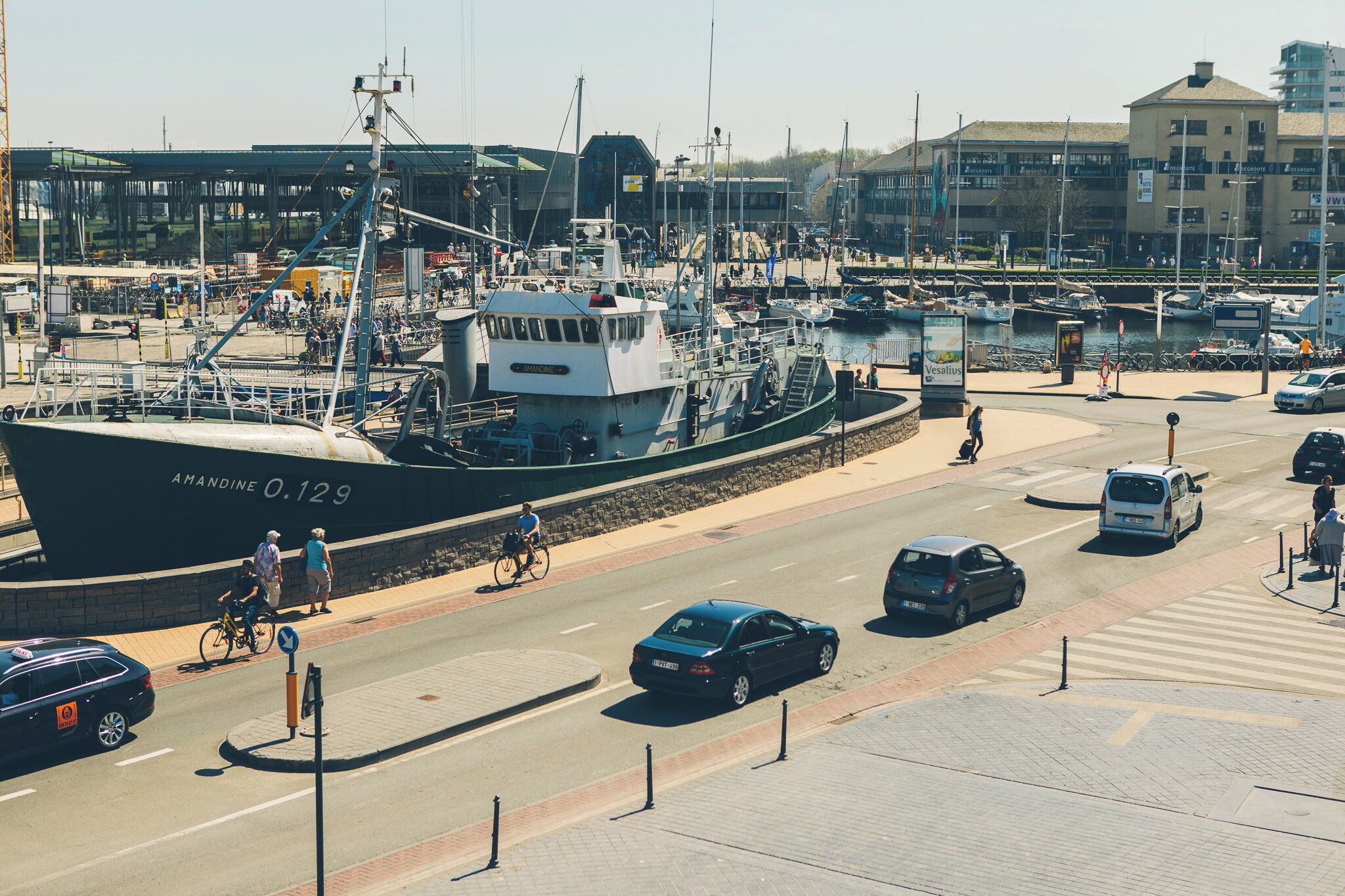 A harbor with a large ship docked, a road with cars and bicycles, and buildings in the background.