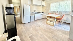 A compact kitchen with white cabinets, a refrigerator, a microwave, and a coffee maker. There is a dining table with two chairs and a window with a view. - Kobayashi Building (Tokyo)
