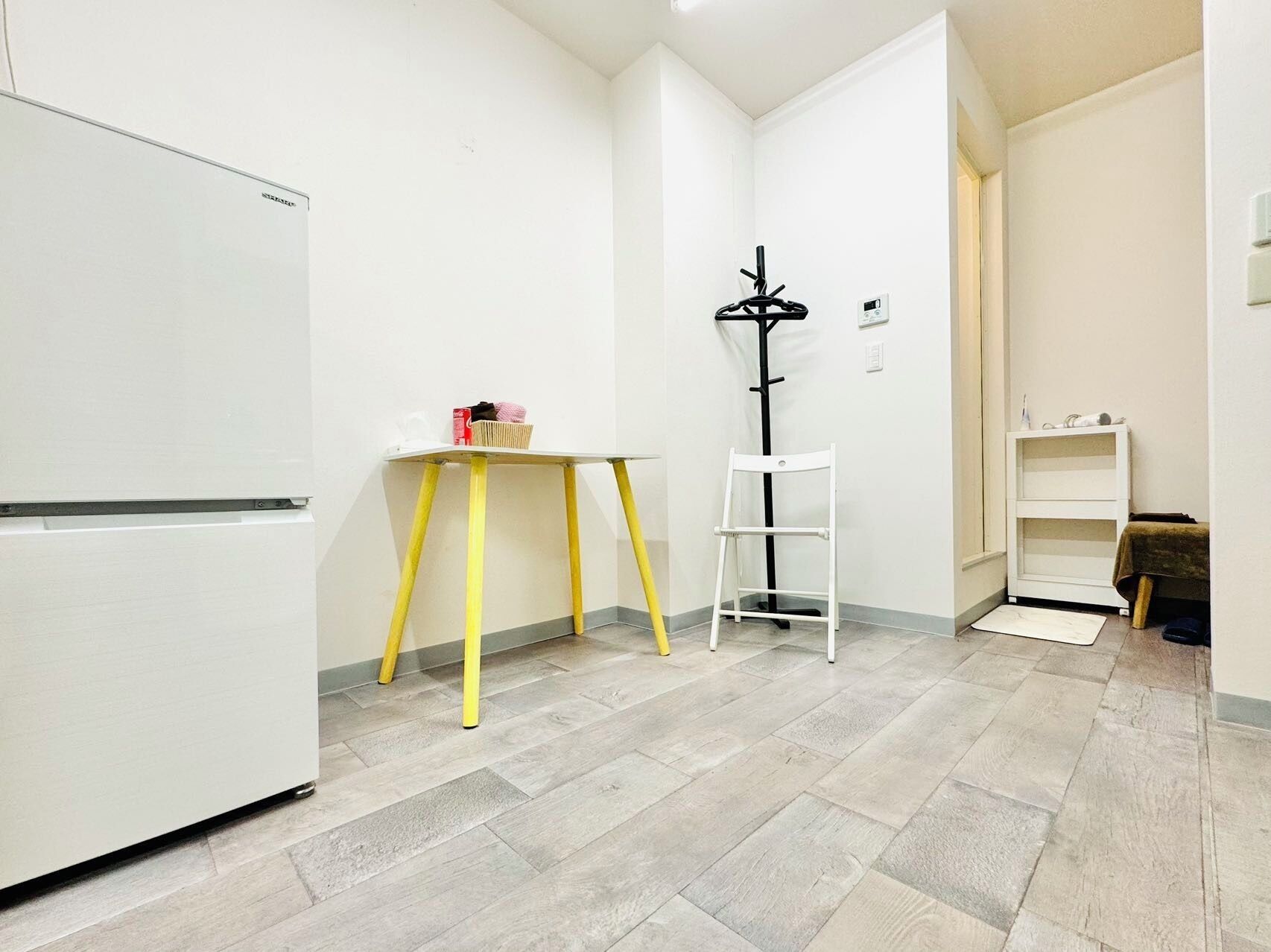 A small, clean room with a white refrigerator, a yellow table, a white shelf, and a black coat rack.