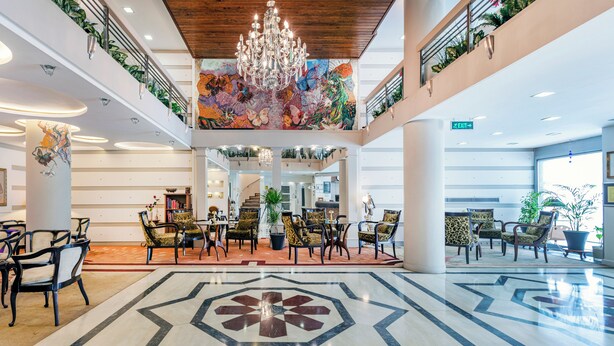 Een ruime lobby met een hoge plafond, een centrale kroonluchter en een kleurrijke muurschildering.