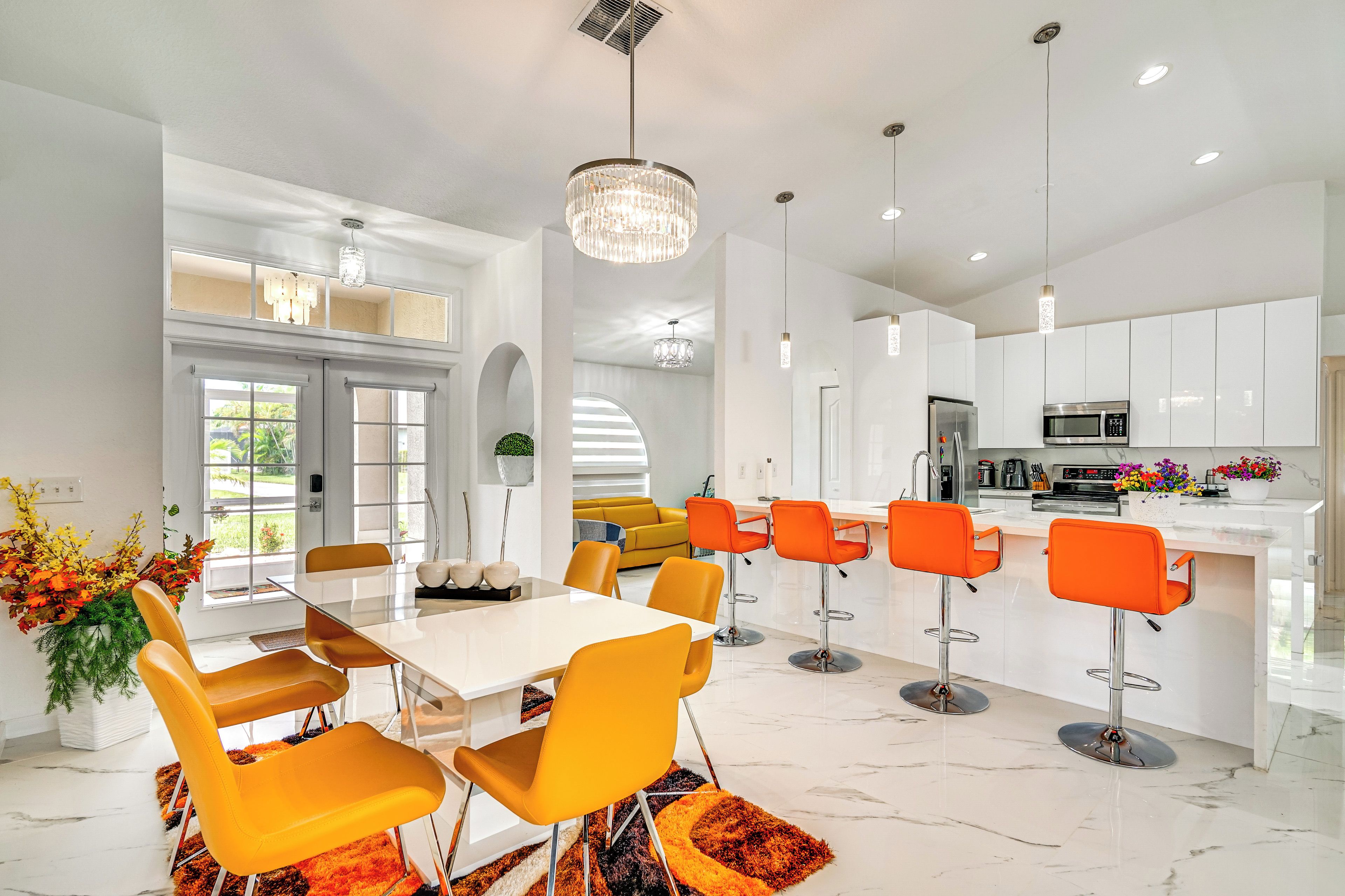 Une cuisine moderne avec des meubles blancs, un îlot central doté de tabourets de bar et un coin repas avec une table blanche et des chaises orange.