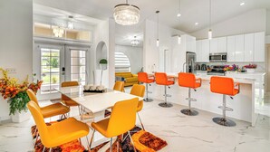 Et moderne køkken med hvide skabe, en central ø med barstole og et spiseområde med et hvidt bord og orange stole.