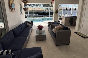 A poolside seating area with two sofas and a coffee table, a view of a swimming pool, and a patio with a stone fireplace.