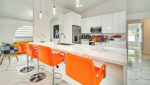 Et moderne køkken med hvide skabe, et rustfrit stål køleskab, en mikrobølgeovn og en vask. Der er tre orange barstole og en hvid marmor bordplade.