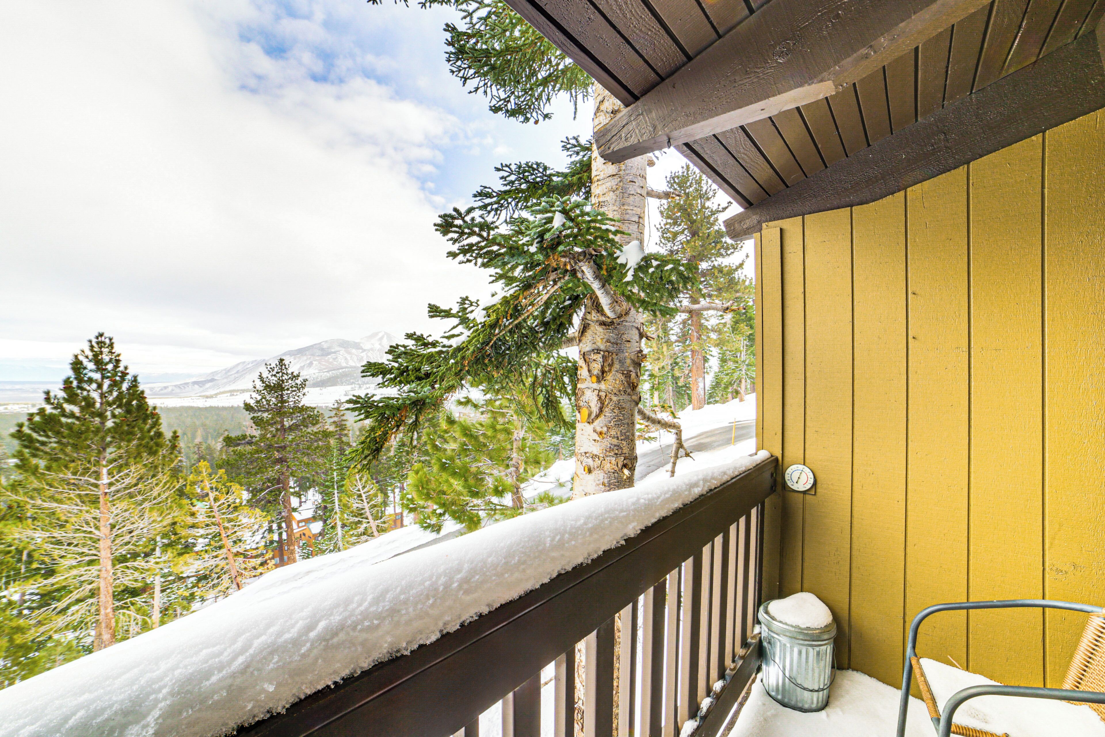 A wooden balcony with snow-covered railings, a wicker chair, and a view of snow-covered trees and mountains.