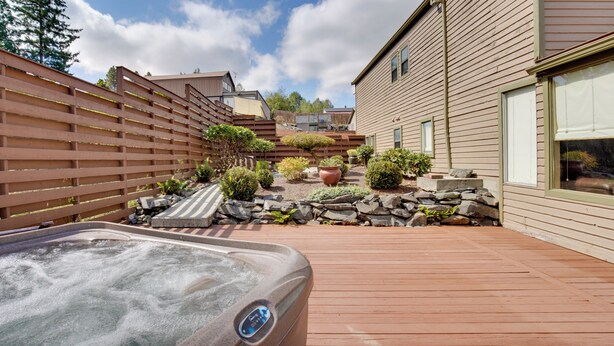Een hottub in een achtertuin met een houten terras, een stenen muur en diverse planten.