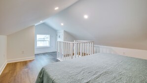 A bedroom with a sloped ceiling, a white crib, a bed with a patterned bedspread, and a view of a house through a window. - Walk to Winery: Holt Home 8 Mi to State Park! (Holt)
