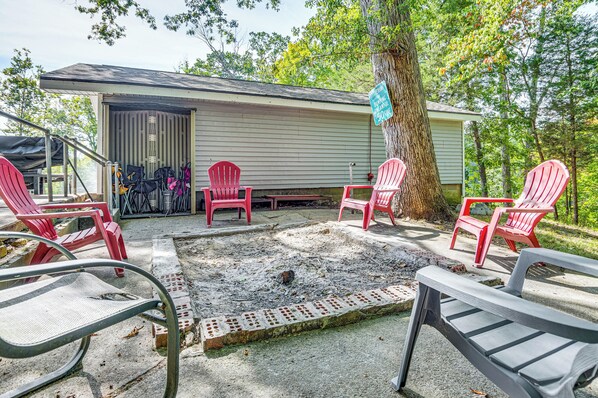 A covered outdoor seating area with red Adirondack chairs and a metal table, surrounded by trees and a building with a garage door.
