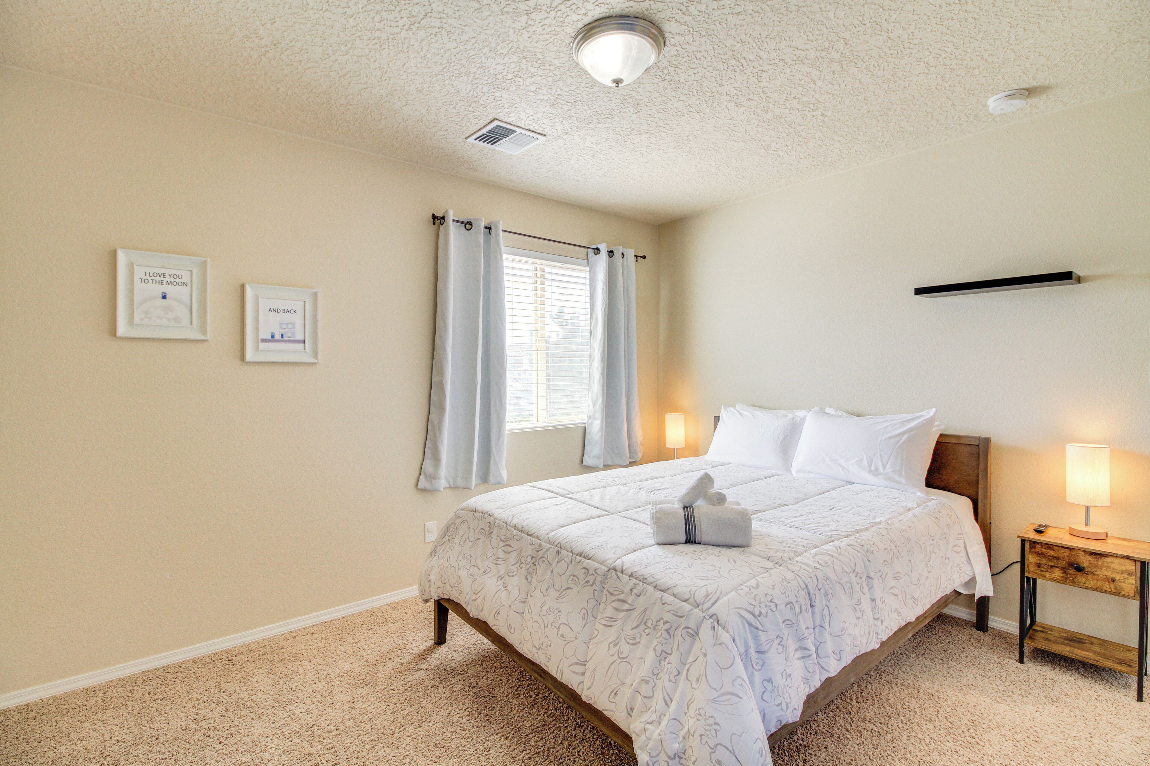 A bedroom with a bed, bedside lamps, a window with curtains, and two framed pictures on the wall.