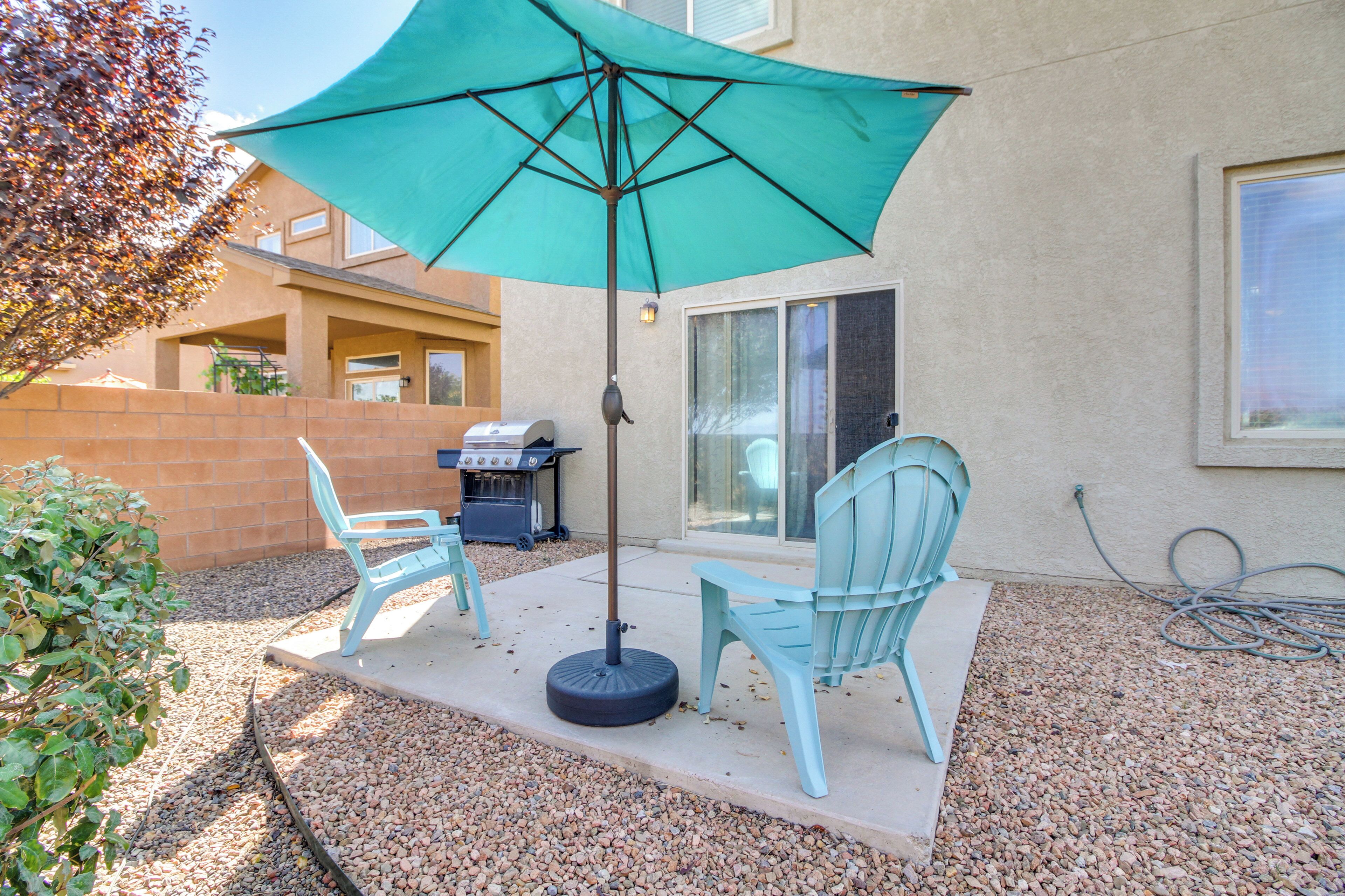 An outdoor patio with a turquoise umbrella, two light blue chairs, and a barbecue grill.