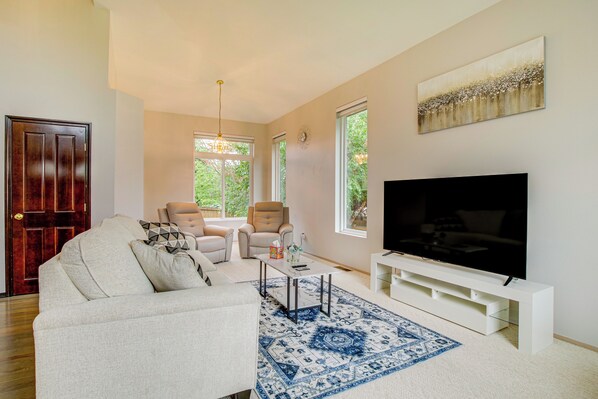 A living room with a sofa, two armchairs, a coffee table, a TV on a stand, and a large window with a view of greenery.
