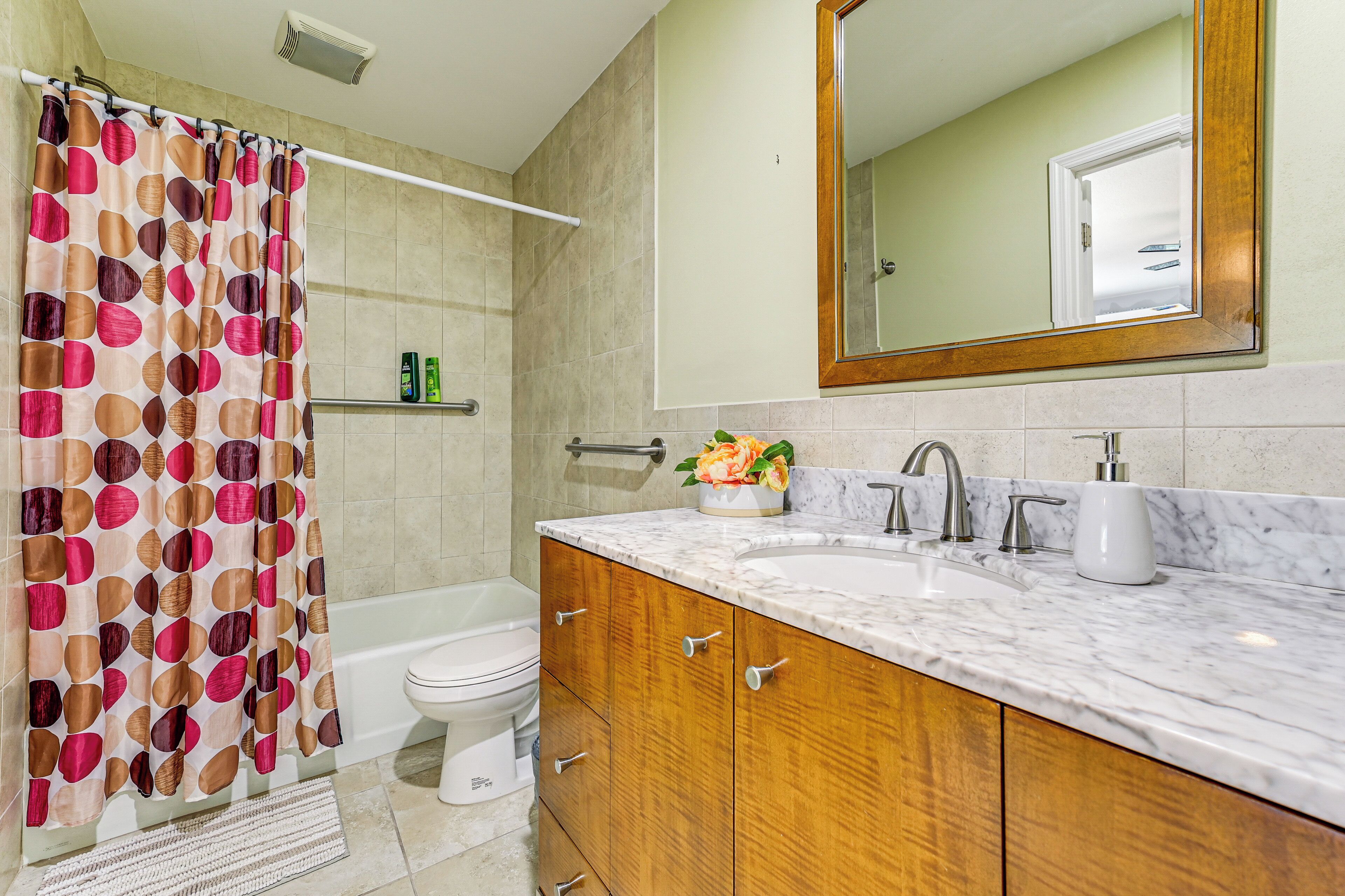 A bathroom with a shower curtain, a bathtub, a toilet, a sink with a marble countertop, wooden cabinets, and a mirror.