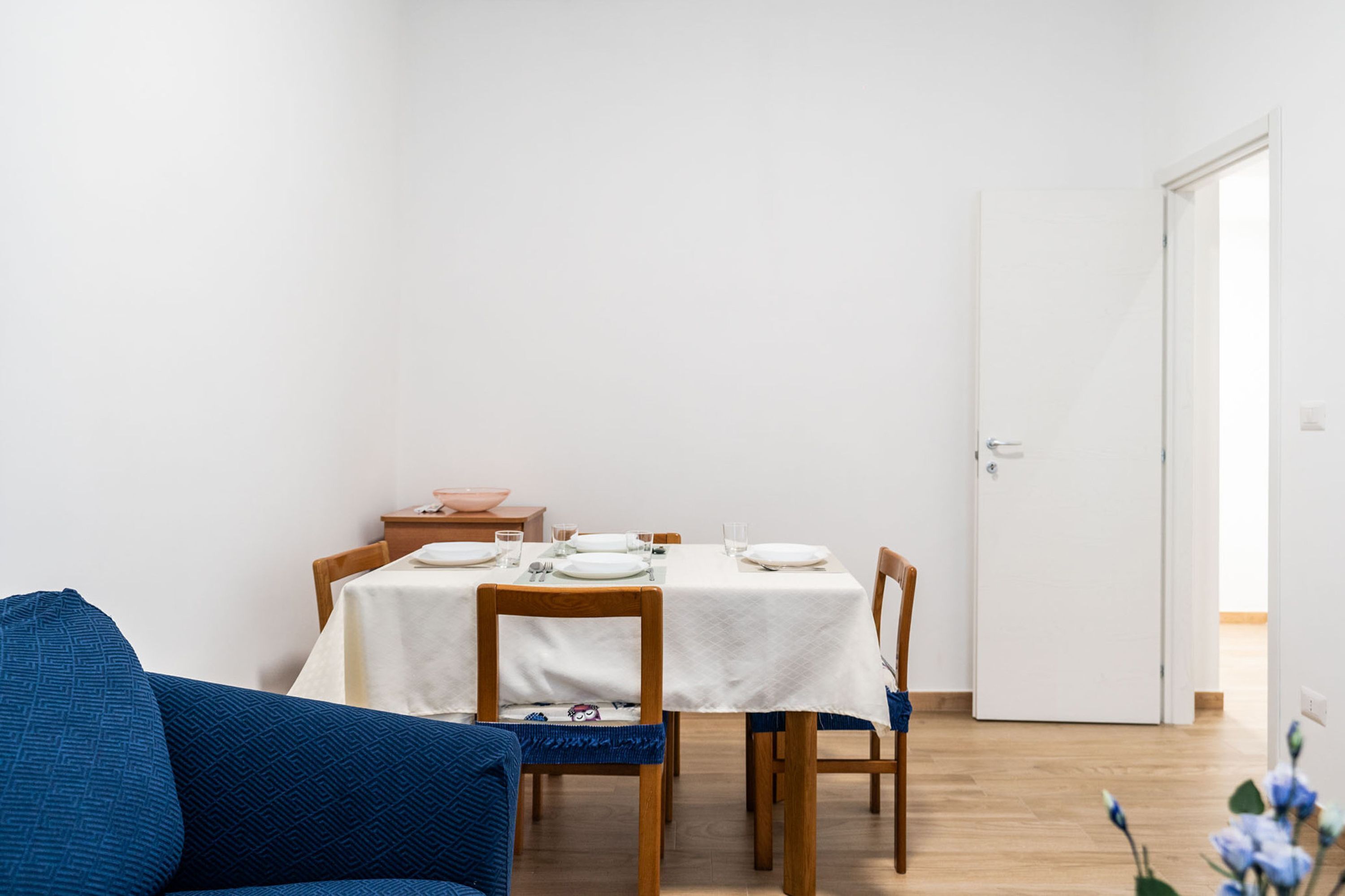 Uma área de jantar com uma mesa posta para quatro pessoas, cadeiras de madeira, um sofá azul e uma porta branca.