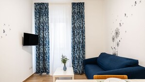 A modern living room with a sofa, a dining table set for two, a flat-screen TV mounted on the wall, and patterned curtains. - The Flower House by Wonderful Italy (Bitritto)
