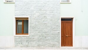 A building facade with a stone wall, a wooden door, and a window with a curtain. - The Flower House by Wonderful Italy (Bitritto)