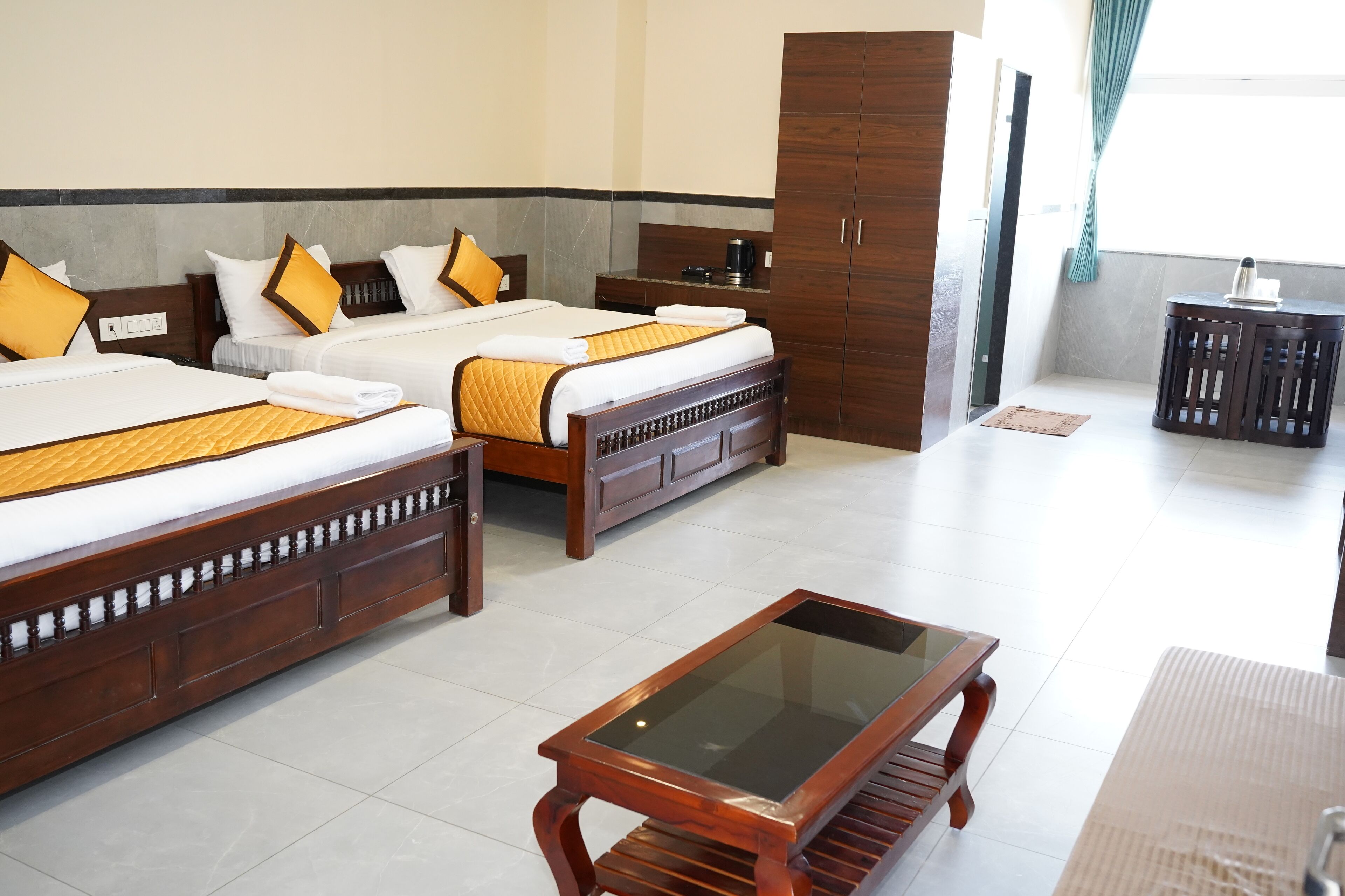 A hotel room with two beds, a wooden wardrobe, a glass-top table, and a small side table.