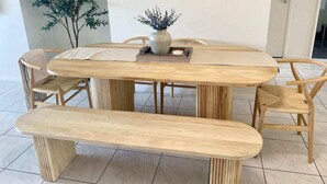 Una mesa de comedor de madera con un banco, sillas tejidas alrededor, un florero con flores y un espejo en la pared.