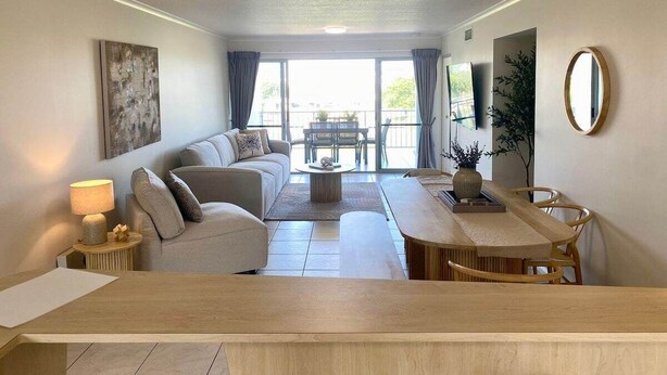 Una sala de estar moderna con un sofá, una mesa de centro, una mesa de comedor y sillas. Hay un ventanal con cortinas, una lámpara en el techo y un cuadro en la pared.