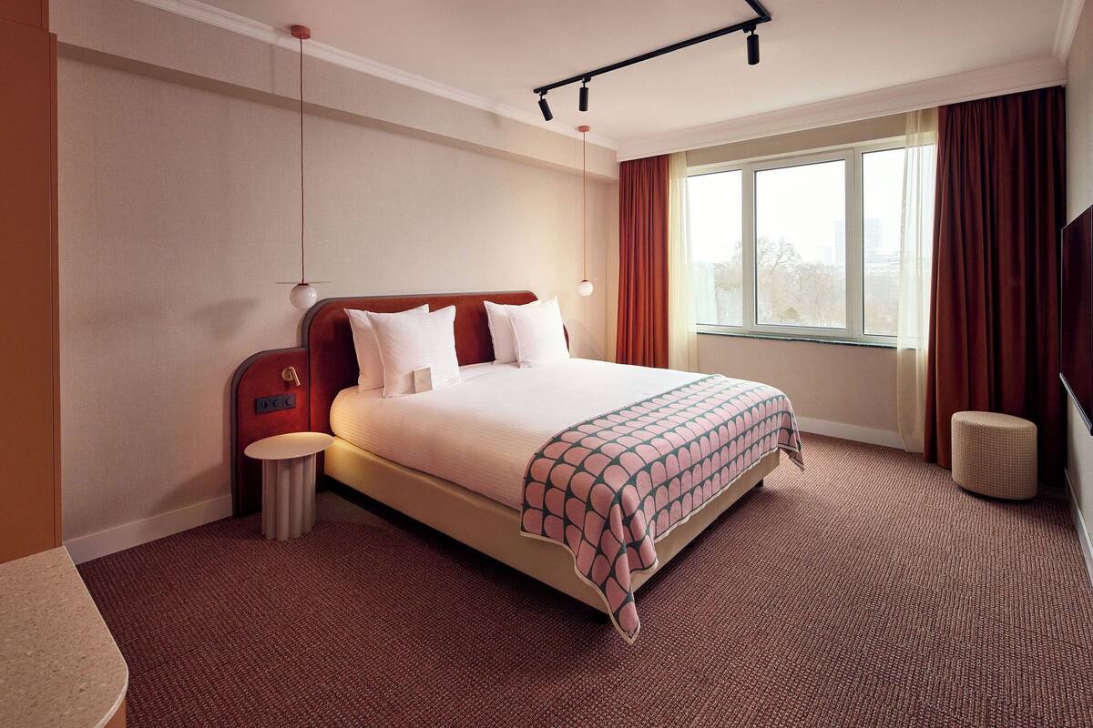 Une chambre d’hôtel avec un grand lit, une table de chevet, une lampe, une fenêtre avec des rideaux et une vue sur la ville.