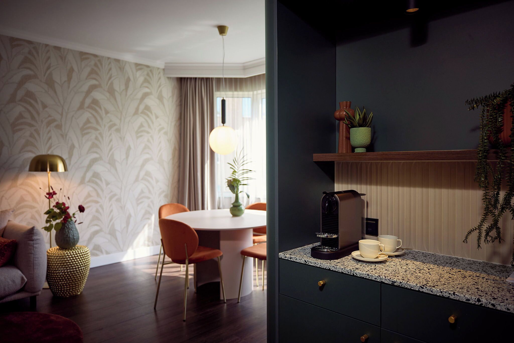A modern living room with a round dining table, orange chairs, a sofa, a lamp, and a coffee machine on the counter.