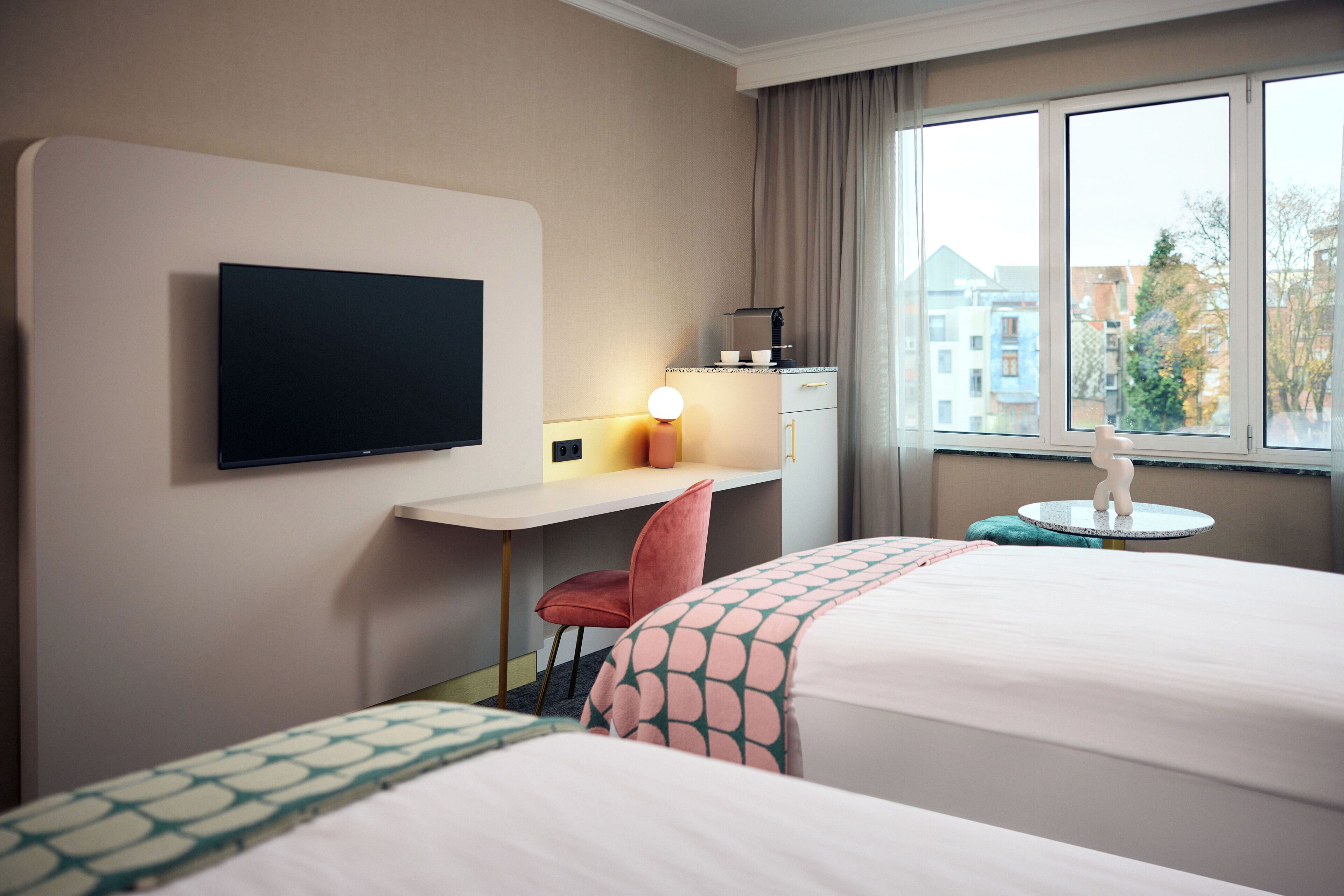 Une chambre d’hôtel avec deux lits, un bureau, une télévision, une lampe et une fenêtre donnant sur des immeubles.
