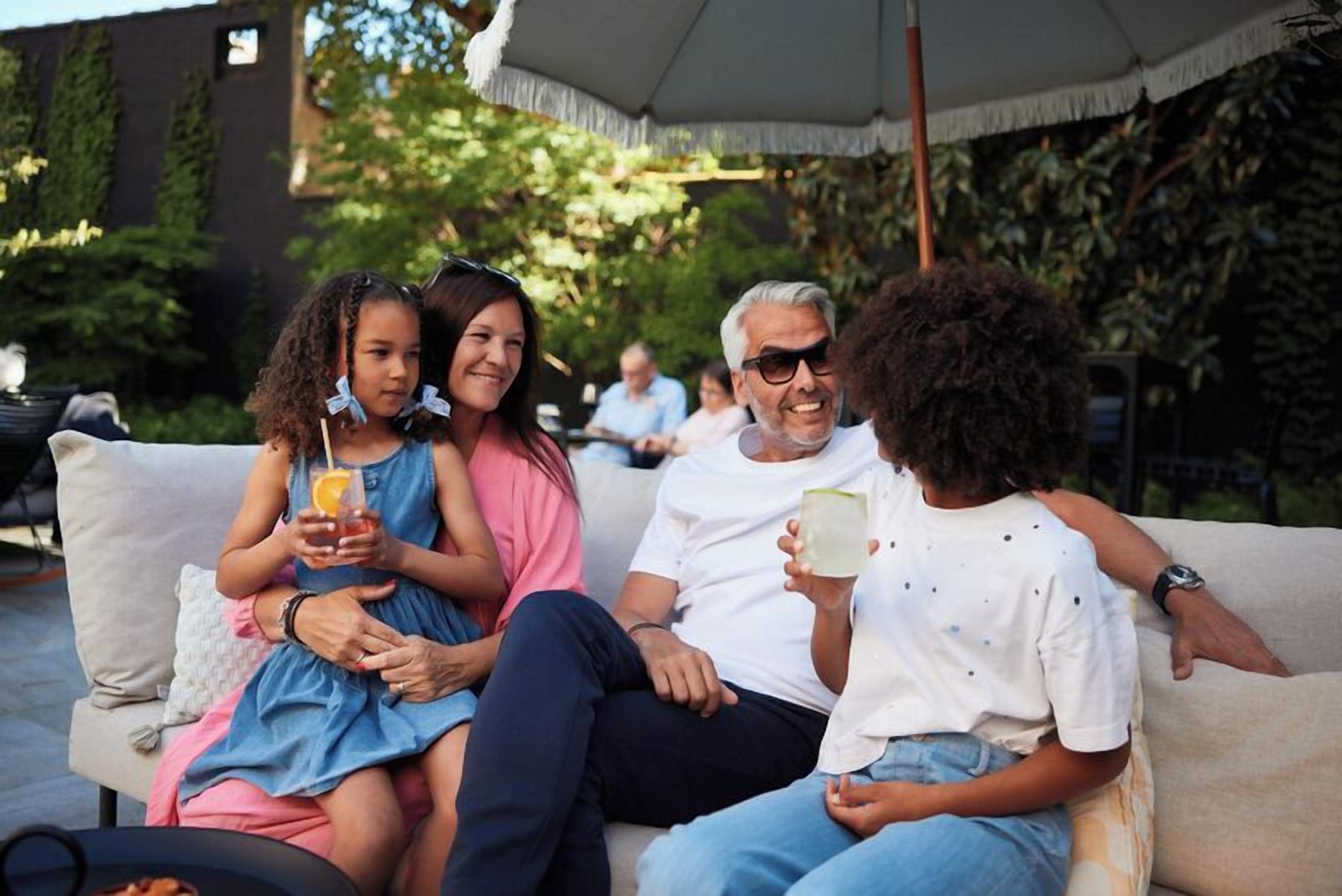 A group of people sitting on a sofa outdoors, with a building and greenery in the background.