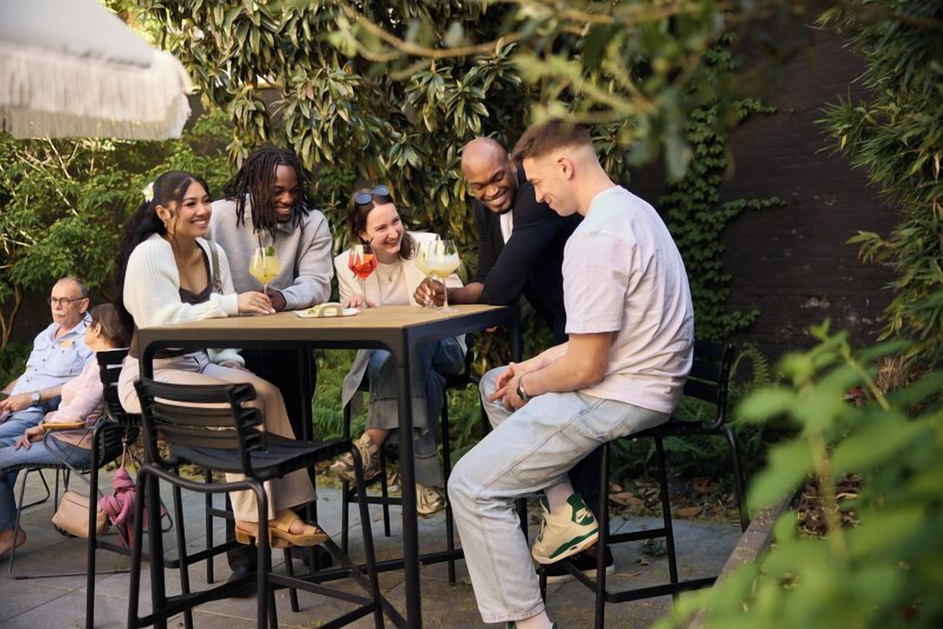 A group of people seated around a table outdoors, with greenery in the background.