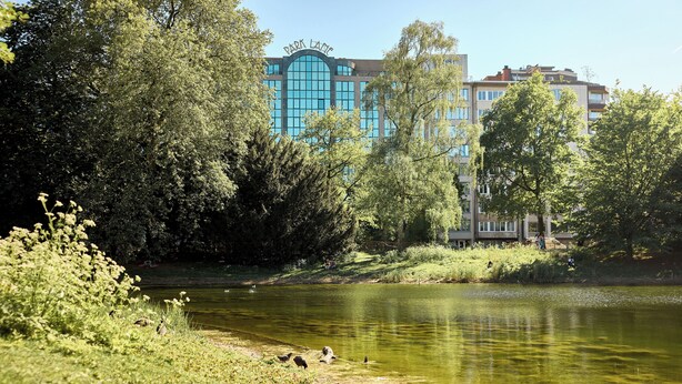 A river with ducks, trees, and a building with 'PARK LATE' on the facade. - Van der Valk Hotel Park Lane Antwerpen (Antwerp)