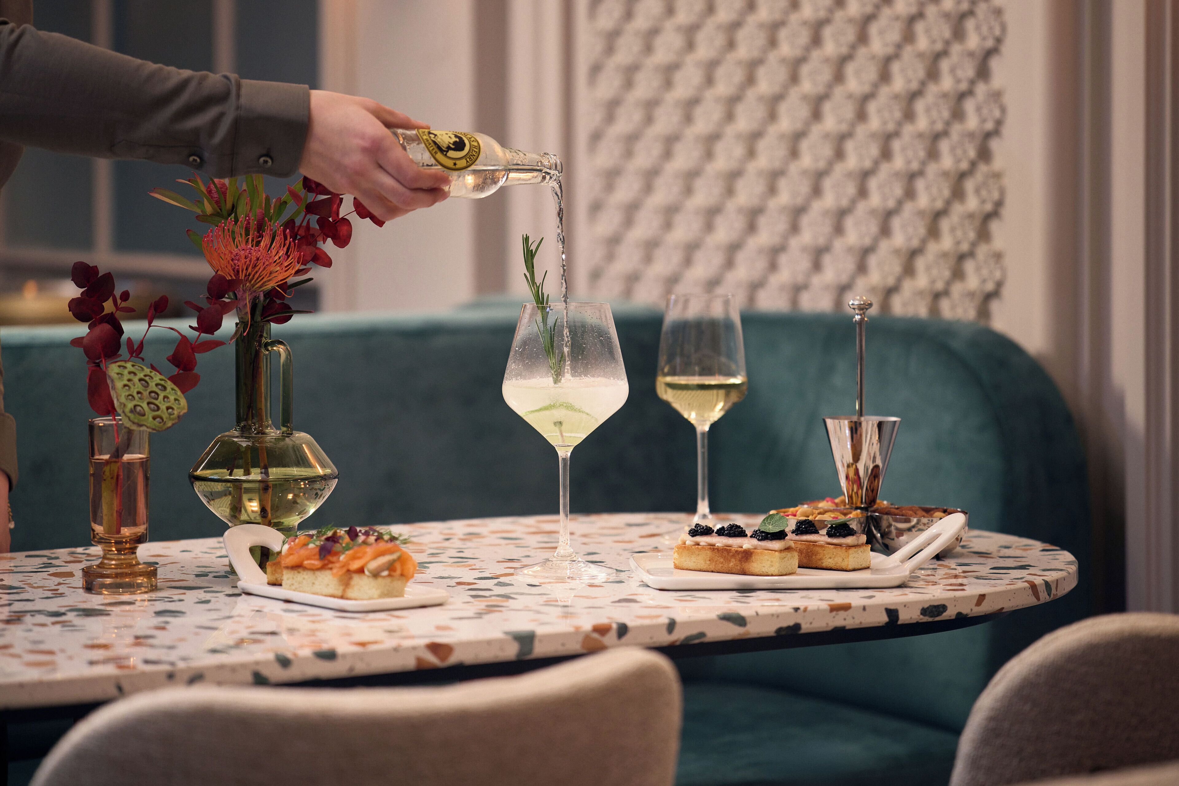 A table with a patterned tablecloth, two glasses of white wine, a bottle of champagne being poured, and a plate with food.