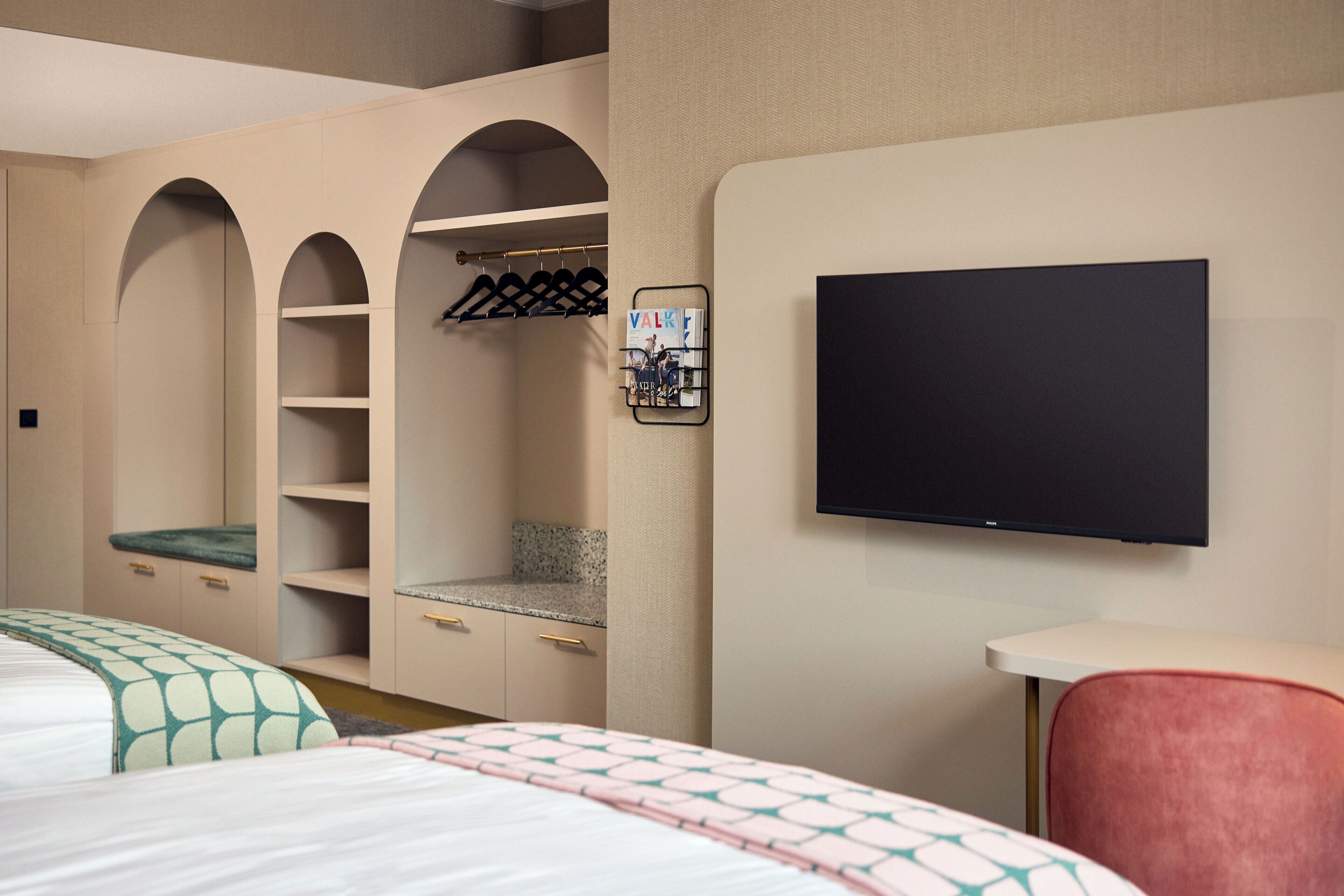 Habitación de hotel con cama, televisor montado en la pared, zona de estar con una silla y armario con estantes y perchero.