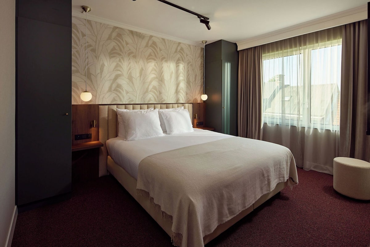 Une chambre d’hôtel avec un grand lit, une table de chevet, une lampe, une fenêtre avec des rideaux et une vue sur l’extérieur.