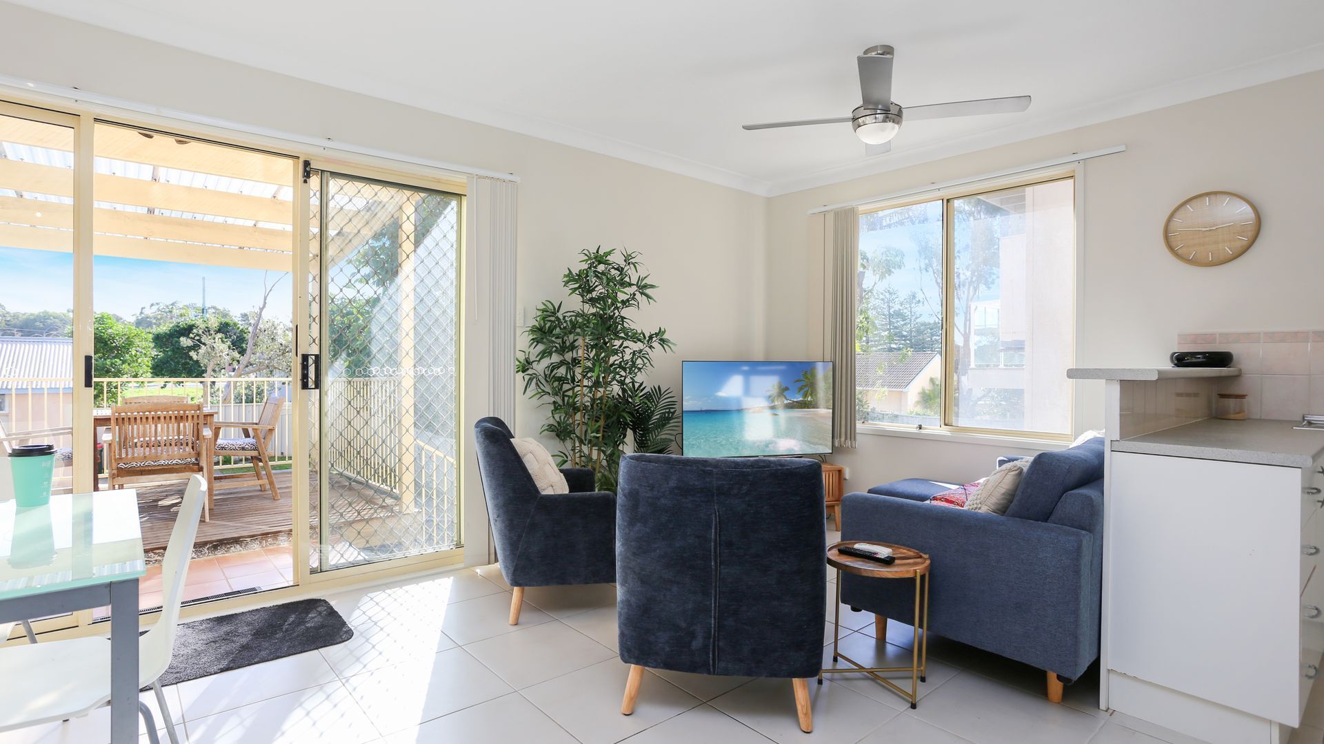 A modern living room with a sliding glass door leading to a balcony, a flat-screen TV, a sofa, and a small kitchen area.
