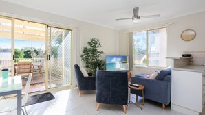En moderne stue med en skydedør, der fører til en balkon, et fladskærms-tv, en sofa og et lille køkkenområde.