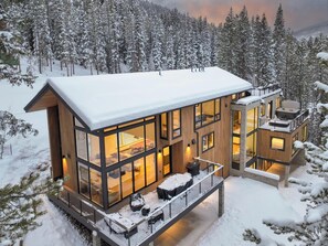 Une cabane moderne en bois, dotée de grandes fenêtres, d’un balcon et d’une terrasse, entourée d’arbres couverts de neige.